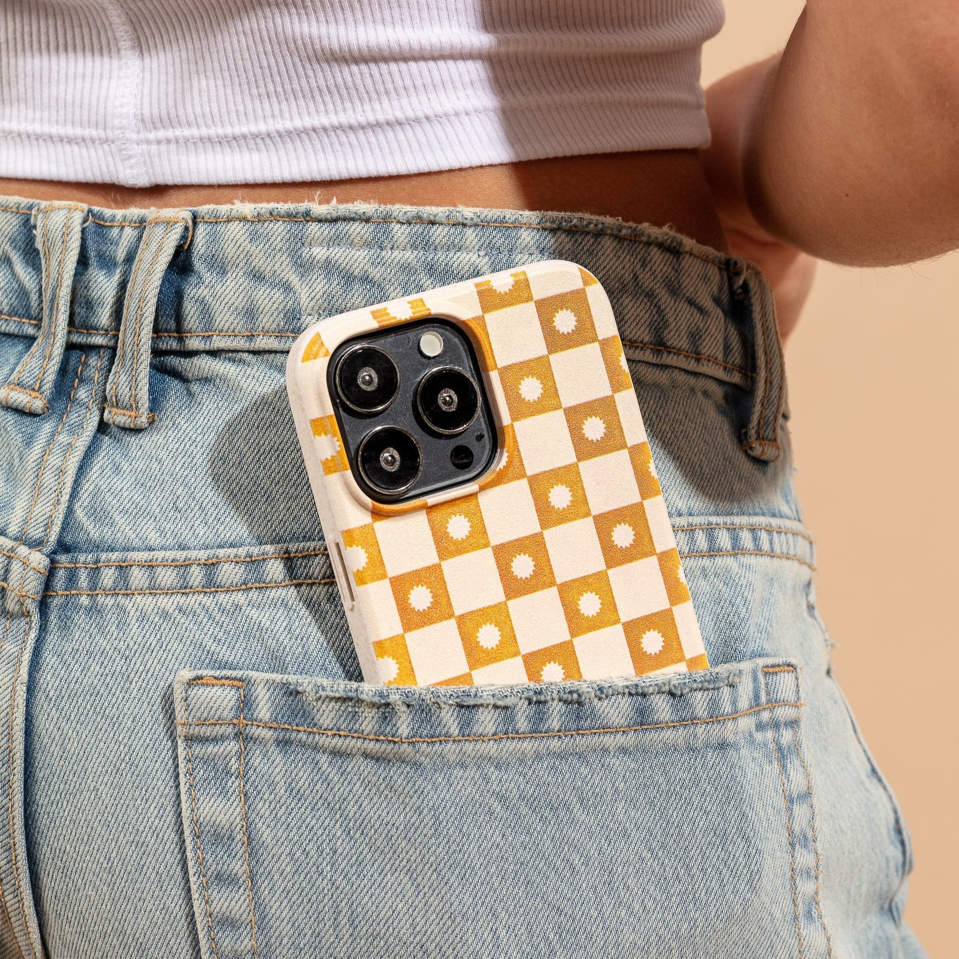 A Star Terracotta Checker Eco-Friendly Phone Case with a yellow and white checkered floral design sticks out of the back pocket of blue jeans, worn by a person in a white crop top.