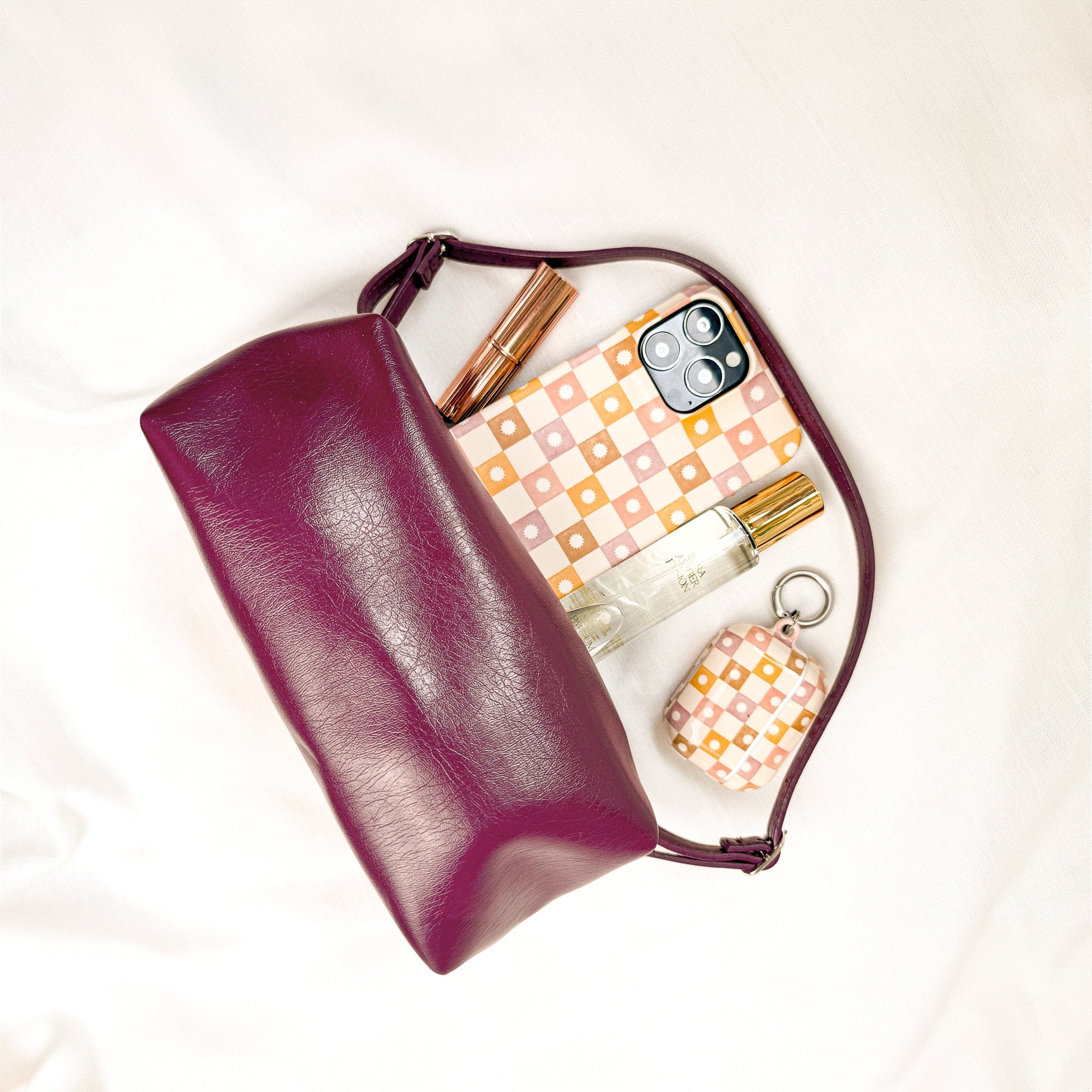 A plum leather pouch on a cream surface sits partially open, revealing a Star Pink Checker Eco-Friendly Phone Case, matching AirPods case, perfume bottle, and two gold makeup items inside.