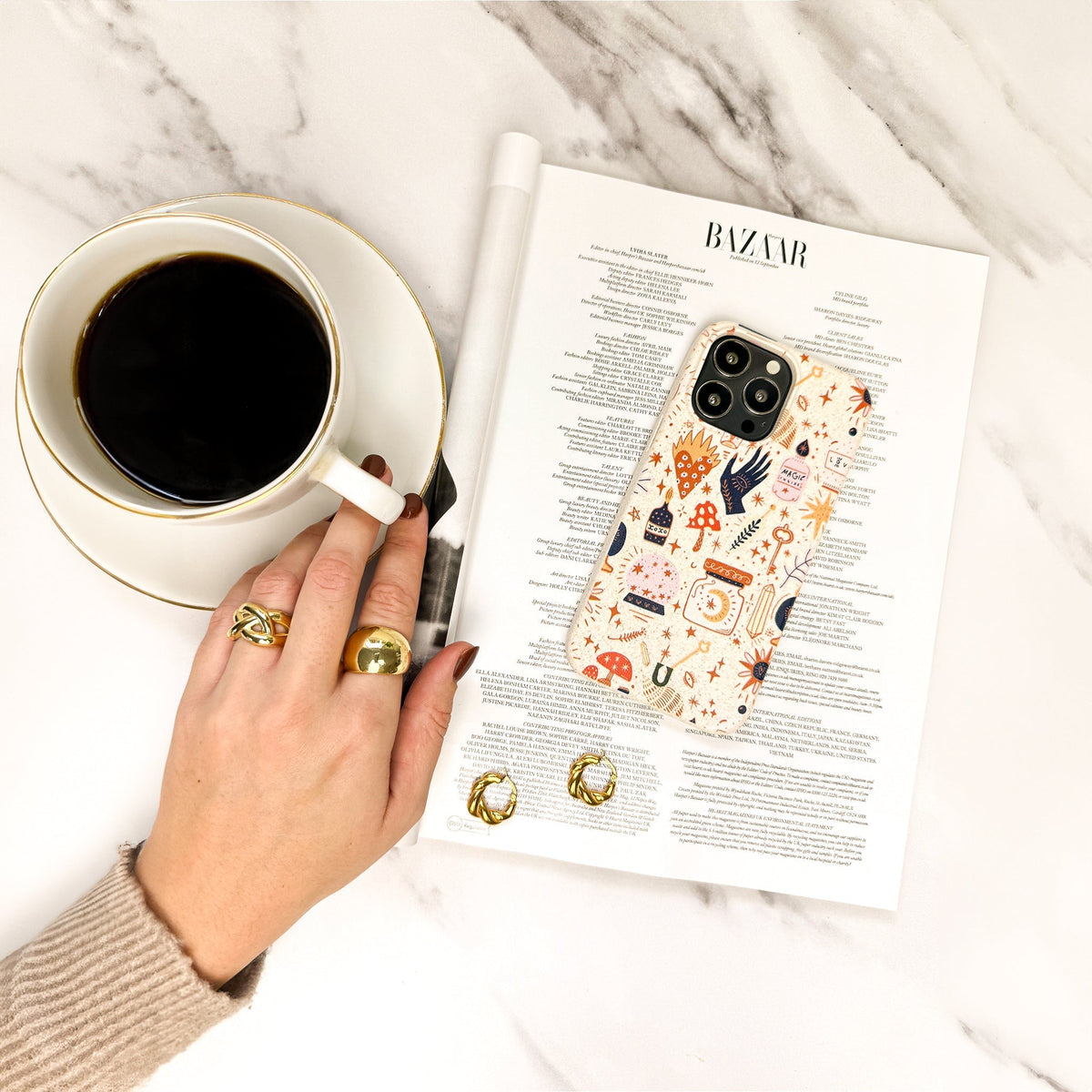 A hand adorned with gold rings rests near a cup of black coffee, a pair of earrings, and a phone with the Love Spell Eco-Friendly Phone Case, all placed on a BAZAAR magazine.