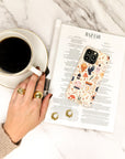 A hand adorned with gold rings rests near a cup of black coffee, a pair of earrings, and a phone with the Love Spell Eco-Friendly Phone Case, all placed on a BAZAAR magazine.