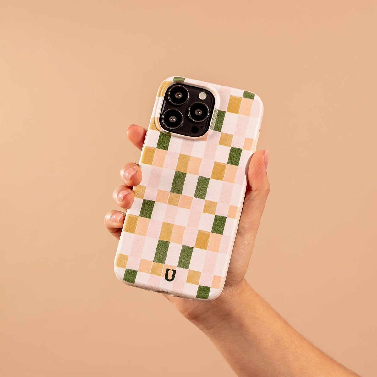 A hand holds a smartphone with the Pink and Green Watercolour Eco-Friendly Phone Case, showcasing a checkered pattern in pastel pink, peach, green, and white tones against a beige background.