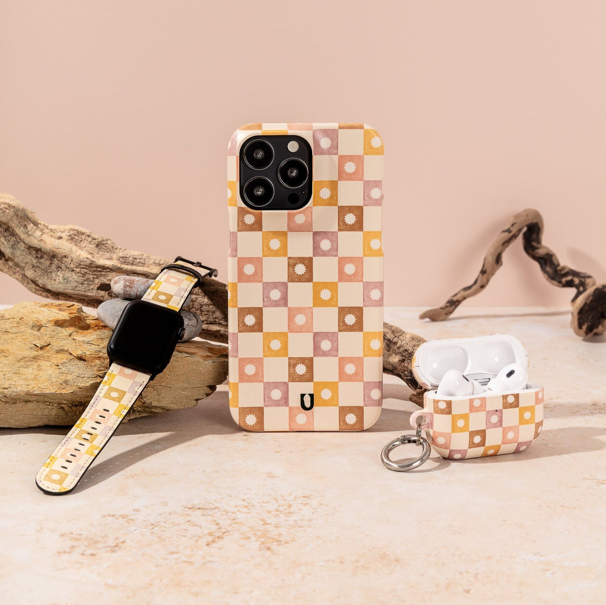 The iPhone, Apple Watch, and AirPods are showcased on a stone surface with driftwood accents in the background, each adorned with the Star Pink Checker Eco-Friendly Phone Case. This case is ideal for anyone looking to express their style while choosing a sustainable option.