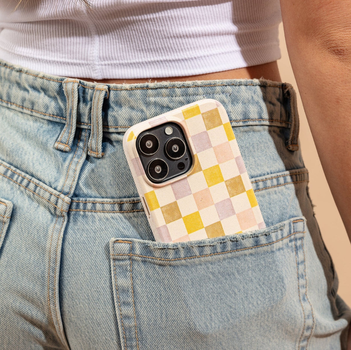 A smartphone with a pink and purple checker eco-friendly phone case peeks out of the back pocket of light blue denim jeans, worn by a person in a white cropped top.