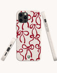 Four views of the Burgundy Bow Ribbons Eco-Friendly Phone Case are shown, featuring a cream background with a red bow pattern. The case includes precise cutouts for buttons, camera, and ports, and is accented by a small green logo near the bottom.