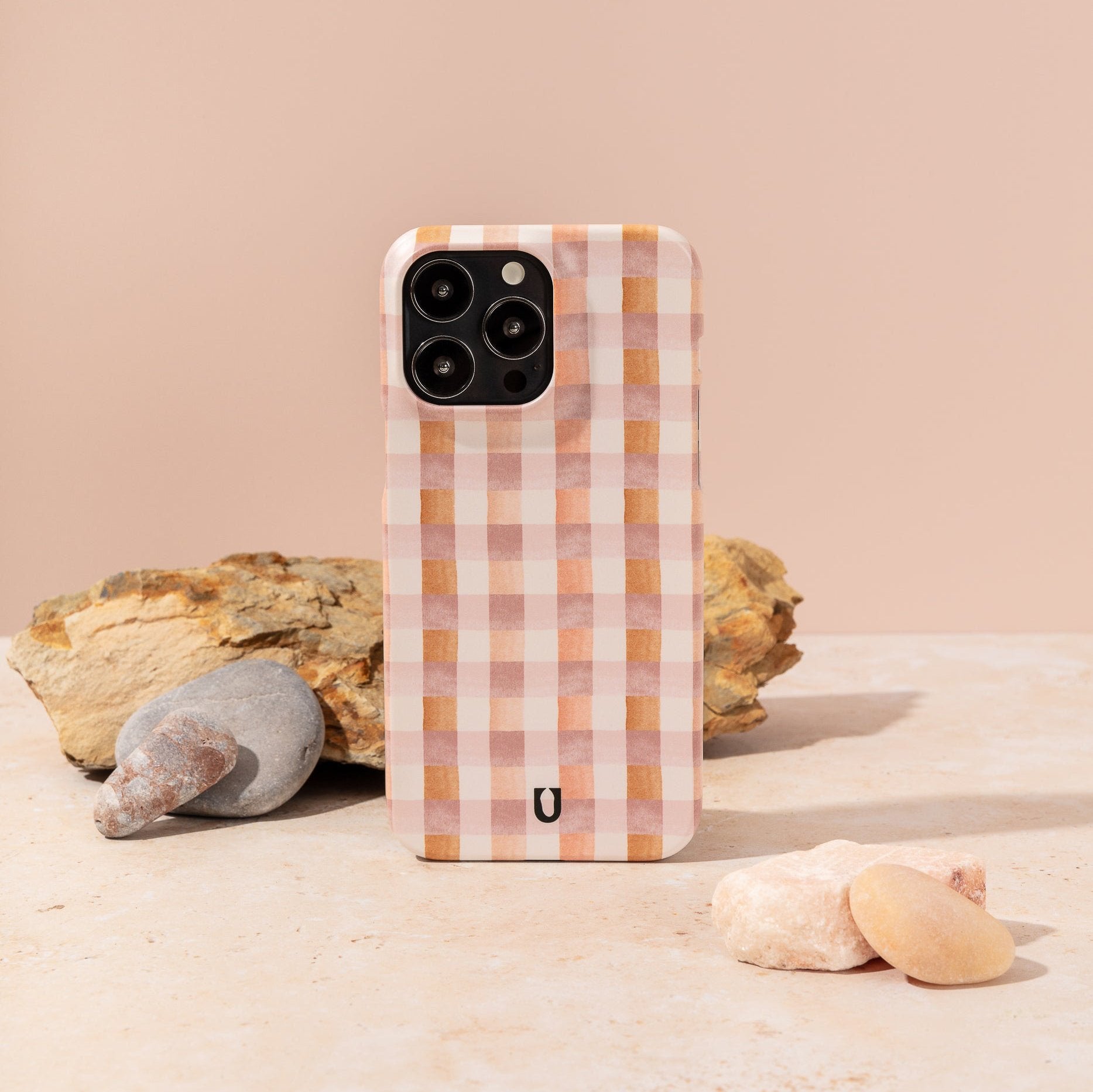 A Pink and Brown Gingham Eco-Friendly Phone Case stands upright on a light surface amid smooth stones and a textured rock, set against a soft peach background.